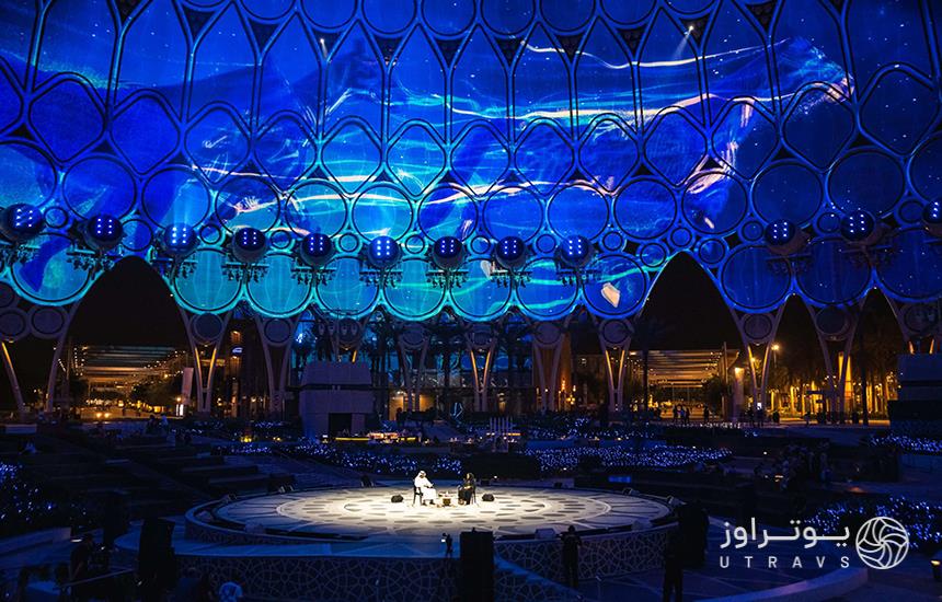 دو نفر نشسته وسط یک سن دایره‌ای‌شکل که پس‌زمینه‌اش با نورهایی به اشکال هندسی تزیین شده در فستیوال آرت دبی
